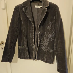 Grå manchesterkavaj från Micha - Snygg grå manchesterkavaj från Micha med broderade blommor på framsidan. Kavajen har en klassisk krage och två fickor framtill. Perfekt för en stilren look.