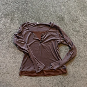 Brun långärmad topp - Snygg brun långärmad topp med knappar framtill. Perfekt för en avslappnad stil. Mjukt och bekvämt material. Har insydd brunt linne som detalj!