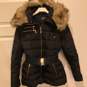 Hollies Avoriaz vinterjacka  - Snygg svart pufferjacka från Hollies med avtagbar fuskpäls på huvan och bälte. Jackan är i princip nyskick, använd enstaka gånger. Den är felfri förutom att på ena fickan fattas dragtapp till blixtlåset (sista bild). Perfekt för kalla dagar med sina praktiska fickor och stilrena detaljer. Priset kan diskuteras!