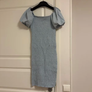 Ljusblå smockklänning från Gina Tricot - Säljer en söt ljusblå smockklänning från Gina Tricot med puffiga kortärmar. Perfekt för en somrig look. Klänningen har en smickrande passform och är tillverkad i ett mjukt material.