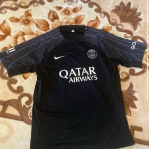 Svart PSG fotbollströja - Snygg svart PSG fotbollströja från Nike med korta ärmar och stilrent mönster på axlarna. Tröjan har klubbens emblem på bröstet och Qatar Airways som sponsorlogga.