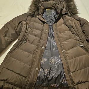 Brun dunjacka från Saki - Snygg brun dunjacka från Saki med avtagbar huva och fuskpäls. Jackan har dragkedja och knappar framtill samt två praktiska fickor med dragkedja. Perfekt för kyliga dagar.