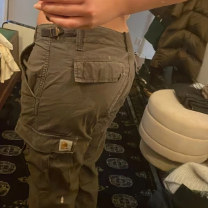 Gröna cargobyxor från Carhartt - Säljer ett par gröna cargobyxor från Carhartt med flera praktiska fickor och en avslappnad passform. Byxorna har en dragkedja och knapp framtill, perfekt för en casual look.