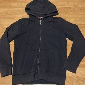 Mörkblå hoodie från GANT - Säljer en mörkblå hoodie från GANT med dragkedja och huva. Tröjan har en broderad logga på bröstet och fickor framtill. 