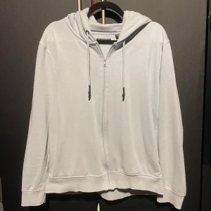 Ljusgrå hoodie från Brave Soul - Säljer en stilren ljusgrå hoodie från Brave Soul. Den har en dragkedja framtill och justerbar huva med snören. Perfekt för en avslappnad look. Det är en unik färg, ljusblå men med mer nyans åt vitt än blå 