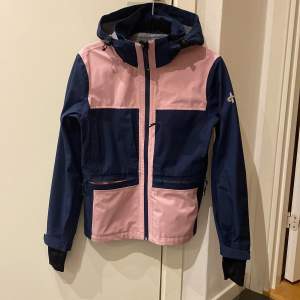 Snygg blå och rosa jacka från Cross Sportswear med dragkedja och avtagbar huva. Perfekt för utomhusaktiviteter med sin funktionella design och praktiska fickor.  Jackan har långa ärmar och en sportig look. 