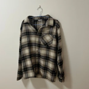 Rutig skjorta - Säljer en snygg rutig skjorta i beige och svart med långa ärmar och klassisk krage. Skjortan har en bröstficka och knappar framtill. Perfekt för en avslappnad stil.