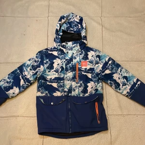 Blå och vit mönstrad skidjacka med huva - Säljer en cool blå och vit mönstrad skidjacka från WARP med huva och dragkedja. 🧥🔵Jackan är storlek 152 i barn. Den har ett unikt bergsmönster och orange detaljer. Luvan går även att ta bort. Hör av er vid minsta fundering!👍😊