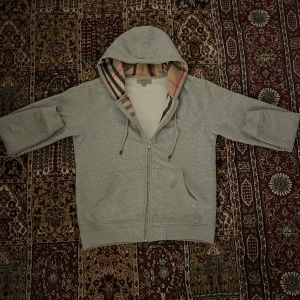 burberry hoodie L sitter som M - Säljer en stilren grå hoodie med dragkedja och en mönstrad insida i huvan. Perfekt för en avslappnad look. Den har långa ärmar och praktiska fickor framtill.