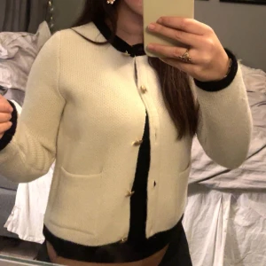 Beige kofta med svarta detaljer - Snygg beige kofta med svarta kanter och dekorativa knappar. Perfekt för en stilren look. Koftan har långa ärmar och två fickor framtill. Dock ganska liten i storlek passar mig som vanligtvis har xs/s