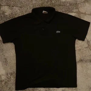 Lacoste tröja - Snygg svart pikétröja från Lacoste med klassisk krage och knappar. Den har det ikoniska krokodilmärket broderat på bröstet. Perfekt för en stilren look. 