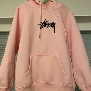 Rosa hoodie från Stüssy - Säljer en snygg rosa hoodie från Stüssy med deras ikoniska logga i svart på bröstet. Hoodien har en stor ficka framtill och justerbar dragsko i huvan. Perfekt för en avslappnad stil!