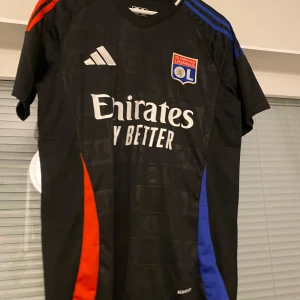 Svart Olympique Lyonnais fotbollströja - Snygg svart fotbollströja från Adidas med Olympique Lyonnais klubbmärke. Tröjan har korta ärmar med blå och röda detaljer på axlarna och sidorna. Perfekt för fotbollsfans som vill visa sitt stöd för laget.
