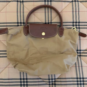 Beige handväska med bruna detaljer - Snygg beige handväska med bruna skinnhandtag och detaljer. Väskan har en praktisk dragkedja och en knapp för extra säkerhet. Perfekt för att bära dina dagliga nödvändigheter med stil.