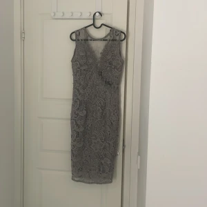 Grå spetsklänning - Elegant grå klänning med vacker spetsdesign. Den har en figurnära passform och är ärmlös, vilket ger en sofistikerad look. Perfekt för festliga tillfällen.