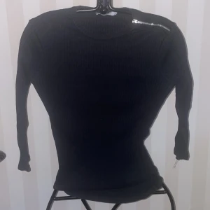 Svart ribbad tröja från Zara - Snygg svart ribbad tröja från Zara med en cool dragkedjedetalj vid axeln. Perfekt för en stilren look. Tröjan har långa ärmar och en figurnära passform.