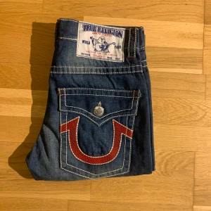 Blå jeans från True Religion - Säljer nu ett par riktigt feta par jeans från True Religion ‼️🔥🔥Jeansen har en straight passform och är dekorerade med kontrastsömmar och knappar samt dess ikoniska bakfickor med röd design ‼️🍾Perfekta för både en avslappnad stil men också om du vill sticka ut lite grann 😉‼️ Jeansen är i väldigt gott skick med rensat små defekter som att lite sömmar lossat lite (bild 5). Passar dig perfekt som är cirka 170 cm‼️Pris kan diskuteras vid snabb affär‼️Tveka inte att höra av er vid frågor!