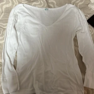 Vit långärmad tröja från Palmers - Säljer en stilren vit långärmad tröja från Palmers. Tröjan har en v-ringad hals med dekorativ spetskant och en liten blomdetalj framtill. Perfekt för en enkel och elegant look.