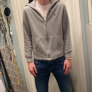 Grå hoodie med dragkedja - Säljer en stilren grå hoodie med dragkedja och huva. Tröjan har långa ärmar och en liten broderad logga på bröstet. Perfekt för en avslappnad look. Ralph Lauren Hoodie i storlek XL barnstorlek, modellen är 190