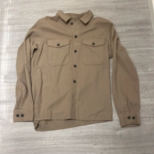 Beige overshirt från Jack & Jones - Snygg beige overshirt från Jack & Jones med långa ärmar och knappar framtill. Perfekt för lager-på-lager-stil. Två bröstfickor med knappdetaljer ger en stilren look.Skick 10/10 då den aldrig har används!!!