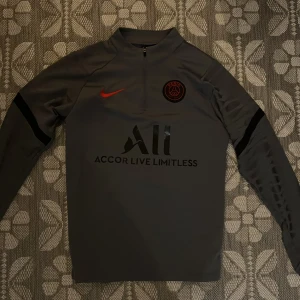 Psg tröja - Säljer en grå träningsjacka från Nike med PSG-logga och texten 'Accor Live Limitless' på framsidan. Jackan har en halv dragkedja och långa ärmar med svarta detaljer. Perfekt för fotbollsträning eller vardagsbruk. Har använt den max 2 gånger och den är helt ny 