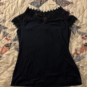 Marinblå topp med spetsdetaljer - Elegant marinblå topp med spetsdetaljer vid axlarna och halsen. Toppen har en smickrande passform och korta ärmar. Perfekt för en stilren look.