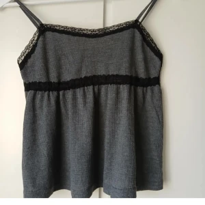 Grå ribbad linnetopp med spetsdetaljer - Snygg grå ribbad linnetopp med svarta spetsdetaljer vid halsringning och midja. Perfekt för en stilren och bekväm look.