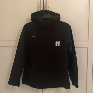 Svart jacka från RevolutionRace - Jackan är helt nytt och har aldrig använts. Snygg svart jacka från RevolutionRace med huva och dragkedja. Perfekt för utomhusaktiviteter med sitt stilrena utseende och praktiska design. Märkeslogga på bröstet och justerbar huva för extra komfort.
