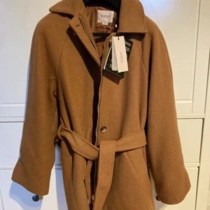 Brun kappa från Soaked in Luxury - Snygg brun kappa i pecan-nyans från Soaked in Luxury. Den har en stilren design med bälte i midjan och dragkedja framtill. Perfekt för höst och vår.