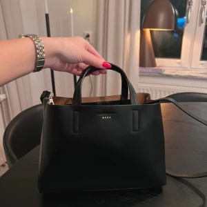 Svart handväska från Wera - Elegant svart handväska från Wera i skinn med stilren design. Väskan har både handtag och en avtagbar axelrem för flexibilitet. Insidan är rymlig med en praktisk ficka för småsaker. Nypris 1200kr.
