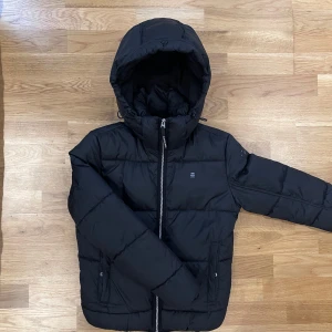 Svart pufferjacka från G-Star RAW - Snygg svart pufferjacka från G-Star RAW med huva och dragkedja. Perfekt för kyliga dagar med sin quiltade design som håller värmen. Använd ett par gånger men i väldigt bra skick