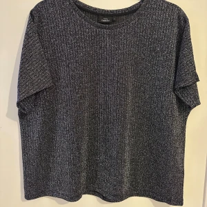 Glittrig topp från Kappahl - Snygg glittrig topp från Kapphal i en mörkgrå nyans. Toppen har en ribbad struktur och korta ärmar. Den är tillverkad i ett syntetmaterial som ger en skimrande effekt.