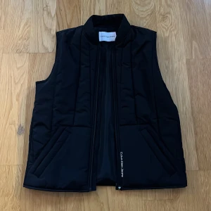 Svart väst från Calvin Klein Jeans - Snygg svart väst från Calvin Klein Jeans med dragkedja framtill och två fickor. Använt två gånger. Nypris 1119kr.