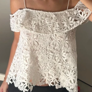 Vit spetsblus med volangdetaljer från Zara - Säljer en söt vit blus i spets med volangdetaljer. Blusen har smala axelband och ett vackert blommönster. Perfekt för sommarens varma dagar.
