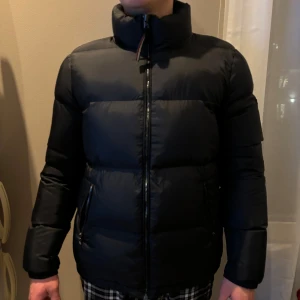 Svart dunjacka från Tommy Hilfiger - Snygg svart dunjacka från Tommy Hilfiger med hög krage och dragkedja. Jackan har en klassisk design med långärmade ärmar och en liten logotyp på ärmen. Oanvänd endast testad då min sambo inte använder den. (Storlek Xl men sitter som en S/M!)