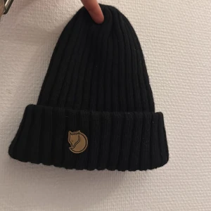 Svart mössa från Fjällräven - Snygg svart ribbad mössa från Fjällräven med ett broderat märke framtill. Perfekt för kyliga dagar och ger en stilren look.