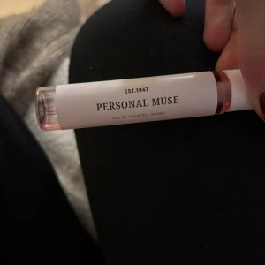 Personal Muse Eau de Toilette - Säljer en elegant Eau de Toilette från Personal Muse. Flaskan är smal och genomskinlig med en vit etikett och rosa lock. Doften är vegansk och flaskan har en stilren design med texten 'EST. 1947'. Perfekt för den som söker en sofistikerad doft.