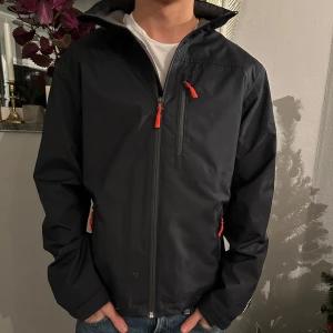 Marinblå jacka från Helly Hansen - Snygg marinblå jacka från Helly Hansen med dragkedja och orange detaljer. Jackan har en hög krage och långa ärmar. Perfekt för en stilren look. Personen på bilden är 180 väger 65 kg! 