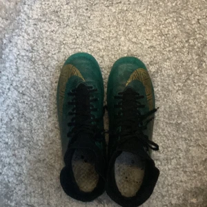 Cristiano Ronaldo Nike fotbollsskor med snörning  - Snygga fotbollsskor från Nike i en grön och svart färgkombination. Skorna har snörning och en socke för att inte grus och gummi pluppar ska störa dig. Perfekta för fotbollsplanen med sina dobbar för bra grepp.