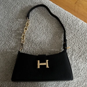 Svart axelväska med gulddetaljer - Snygg svart axelväska med en elegant guldkedja och ett stort H-spänne framtill. Väskan har en stilren design och är perfekt för att lyfta din outfit. Den är helt ny och aldrig använd. Köpte väskan i en butik i Dubai💞💞