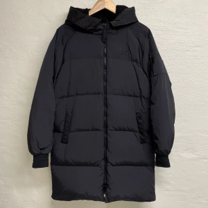 Svart dunjacka från Adidas - Snygg svart dunjacka från Adidas med huva och dragkedja. Jackan har långa ärmar och en broderad logga på bröstet. Perfekt för kalla dagar med sin varma och vadderade design.