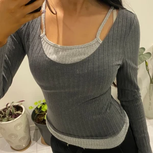 Grå ribbad tröja med dubbla axelband - Snygg grå ribbad tröja med unika dubbla axelband och långärmad design. Perfekt för en stilren look med en twist. Passar bra till både jeans och kjol.