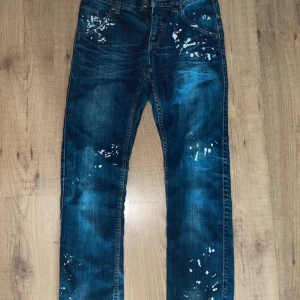 Blå jeans med färgstänk från We Tweens - Snygga blå jeans från We Tweens med unika vita färgstänk för en cool look. De har en straight passform och klassisk femficksdesign. Perfekta för att ge din outfit en kreativ touch.