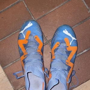 Blå och orange fotbollsskor från Puma - Snygga blå fotbollsskor från Puma med orange detaljer och snörning. Perfekta för fotbollsplanen med en stilren design och bra grepp.