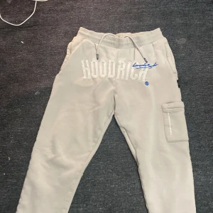Beige mjukisbyxor från Hoodrich - Säljer ett par beige mjukisbyxor från Hoodrich med broderad logga i vitt och blått. Byxorna har snörning i midjan och en praktisk ficka på sidan. Perfekta för en avslappnad stil.