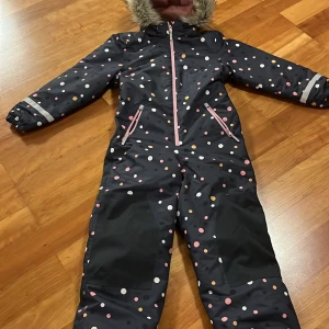 Mönstrad overall från H&M - Säljer en härlig mörkblå overall från H&M med färgglada prickar i rosa, vitt och gult. Den har en mjuk och varm fuskpäls runt huvan samt vackra rosa dragkedjor. En perfekt och stilig overall för kyliga dagar!