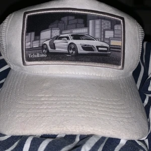 Vit keps med bilmotiv från VeluRoso - Snygg vit keps från VeluRoso med ett coolt bilmotiv på framsidan. Kepsen har ett nätmaterial på sidorna och baksidan för extra ventilation. Perfekt för bilentusiaster! 75/100 gjorda. Vunnen på dreamhack 2023 av en youtuber.