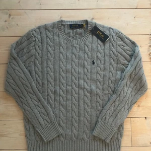 Grå kabelstickad tröja från Polo Ralph Lauren - Säljer en stilren grå kabelstickad tröja från Polo Ralph Lauren. Tröjan har en klassisk rund hals och det ikoniska logotypbroderiet på bröstet. Perfekt för kyligare dagar. 💕