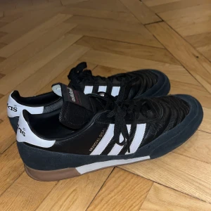 Adidas sneakers med svarta och vita detaljer - Snygga Adidas sneakers i svart med vita ränder och detaljer. Skorna har en klassisk design med snörning och en brun sula. Perfekta för en sportig look.