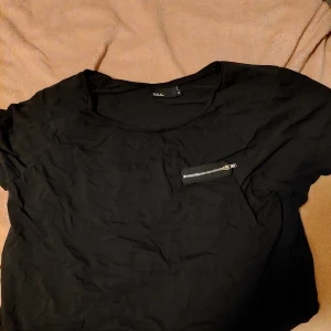 Svart topp med dragkedja från Zoul - Säljer en svart topp från Zoul med en cool dragkedjedetalj på bröstet. Perfekt för en stilren look. Toppen har en rund halsringning och korta ärmar.